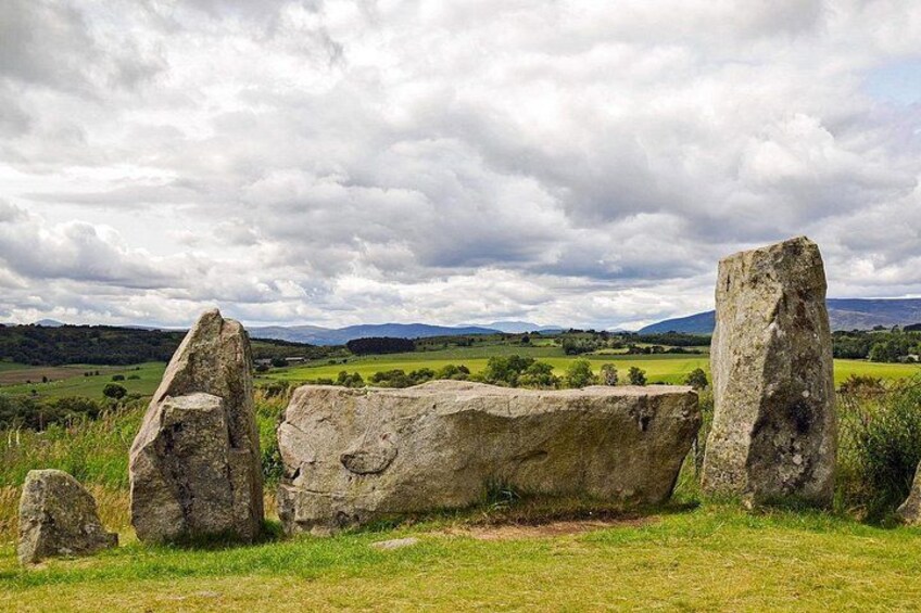 A stone circle with large upright stones and a flat stone in the center, surrounded by grassy fields and distant hills under a cloudy sky. 3