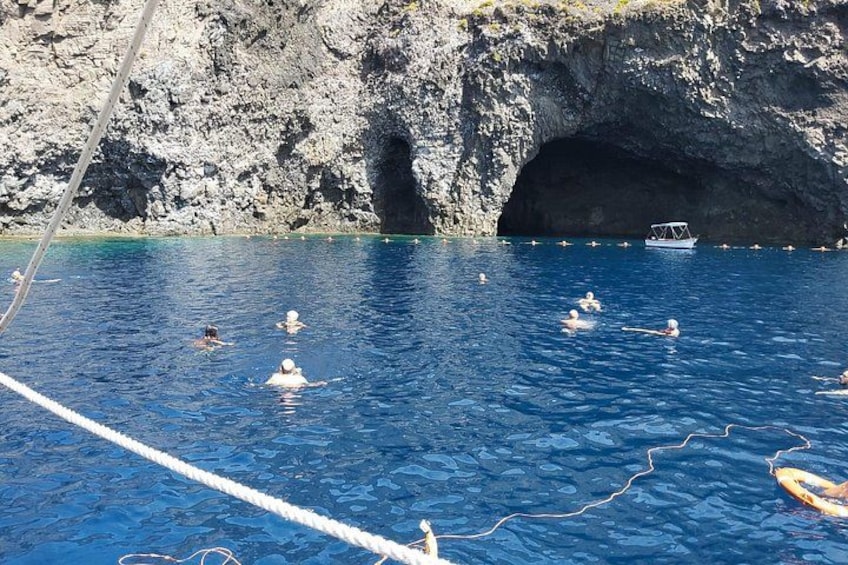 A group of people swimming in a clear blue sea with a rocky cliff and a boat in the background. 3
