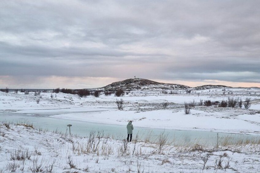 A snowy landscape with a frozen lake, a person standing by the lake, and a hill in the background. 2