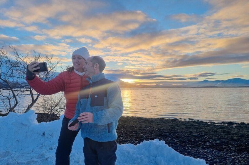 Two people taking a selfie by a snowy lakeside with a sunset in the background. 1