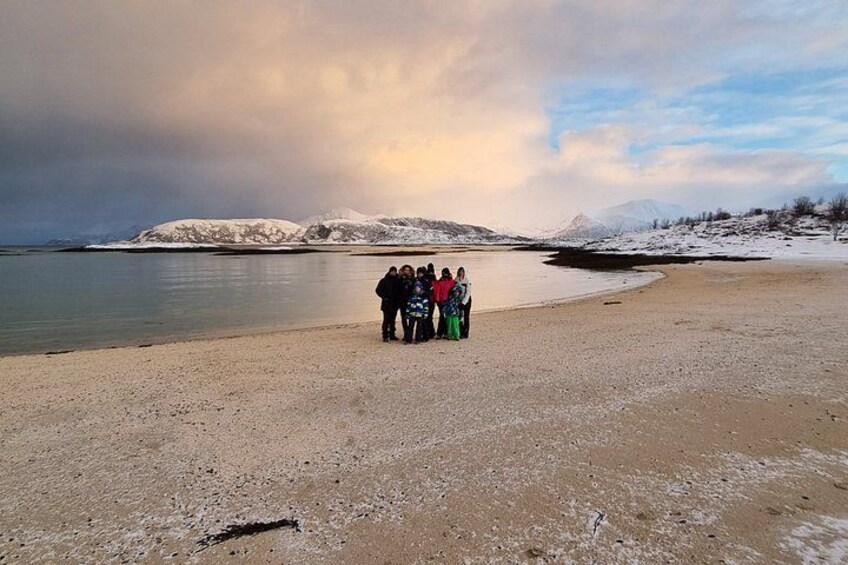 A group of people standing on a pebbly beach with snow-covered mountains in the background. 3