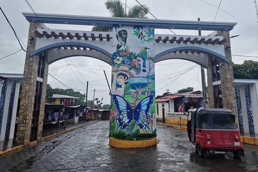 Un mural colorido en un portón, un vehículo rojo estacionado a un lado, y una calle mojada. 2