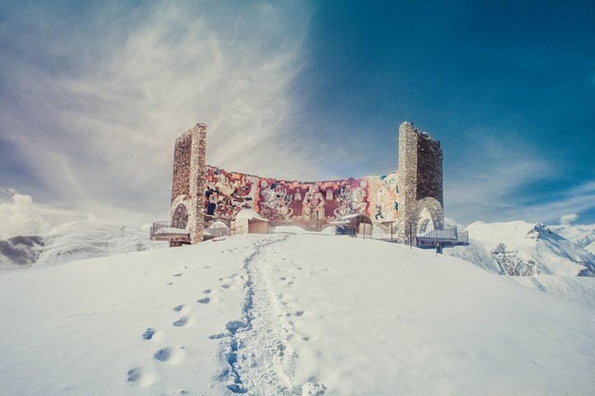 A snow-covered landscape with a historic building featuring colorful murals, a clear sky, and a snow-covered path leading to the structure. 2