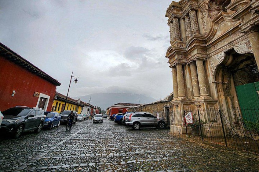 A cobblestone street lined with parked cars and buildings, including a large ornate structure on the right. 1