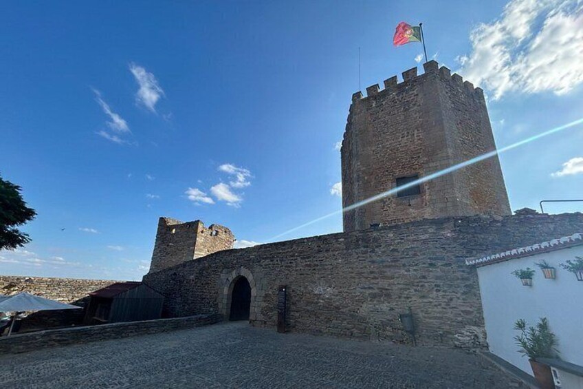 Et historisk stenkastel med flag på taget, en stor buegang og en klar blå himmel. 1