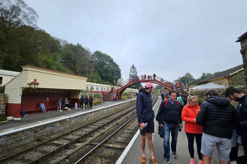A train station platform with people waiting, a train track, and a red bridge. 2