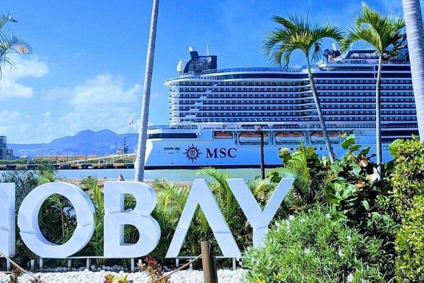 A cruise ship docked at a port with the MSC logo, palm trees, and the word 'IBAY' in the foreground. 3