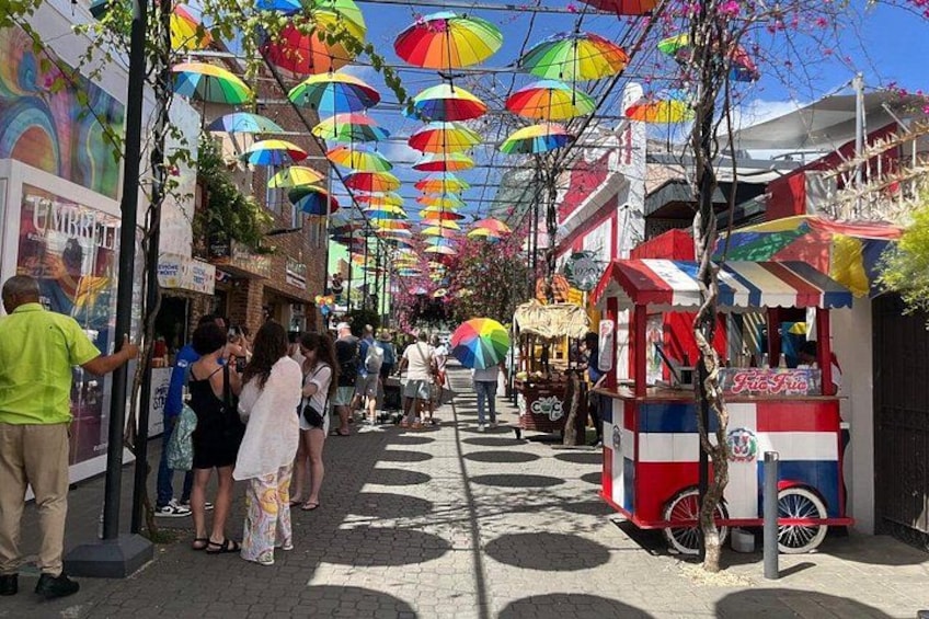 A street lined with colorful umbrellas, a food cart, and people walking. 2