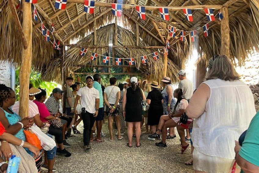A group of people gathered under a thatched roof structure with flags. 1