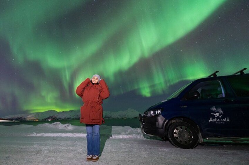 A person in a red coat standing next to a blue car with a logo, under a night sky with green aurora. 3