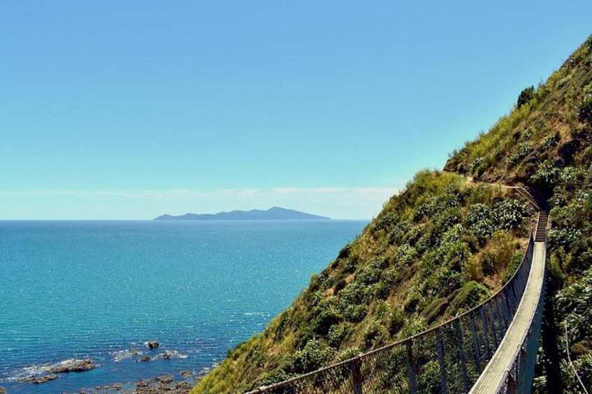 A coastal road with a guardrail, a clear blue sea, and a hilly landscape. 1