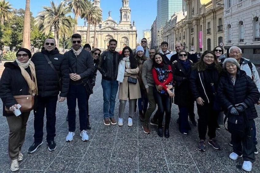Un grupo de personas de pie en una plaza con palmeras y un edificio histórico al fondo. 2