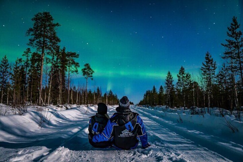 Two people sitting on a snow-covered path, facing the northern lights. 4