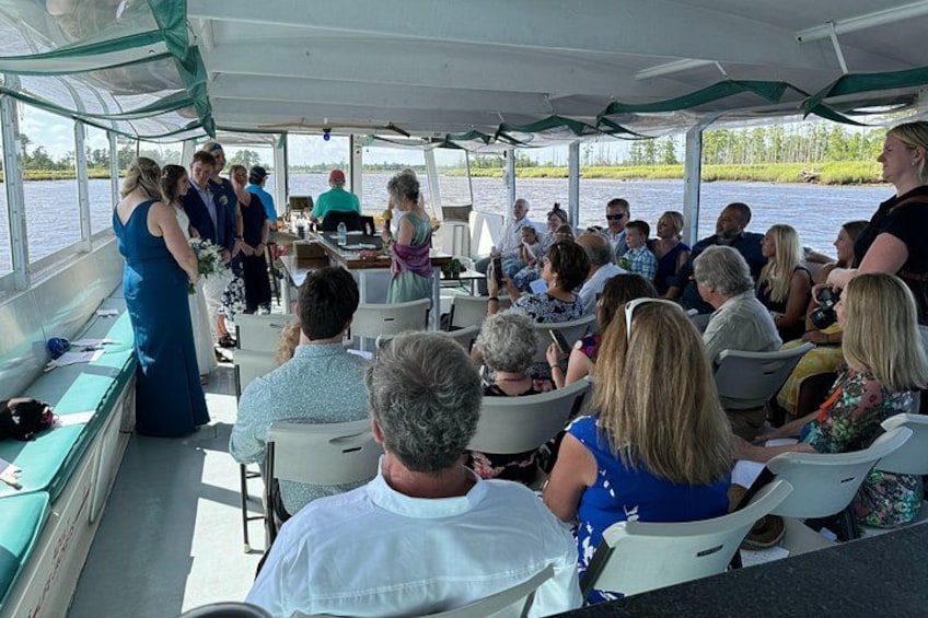 A group of people on a boat, some standing and some seated, with a river in the background. 2