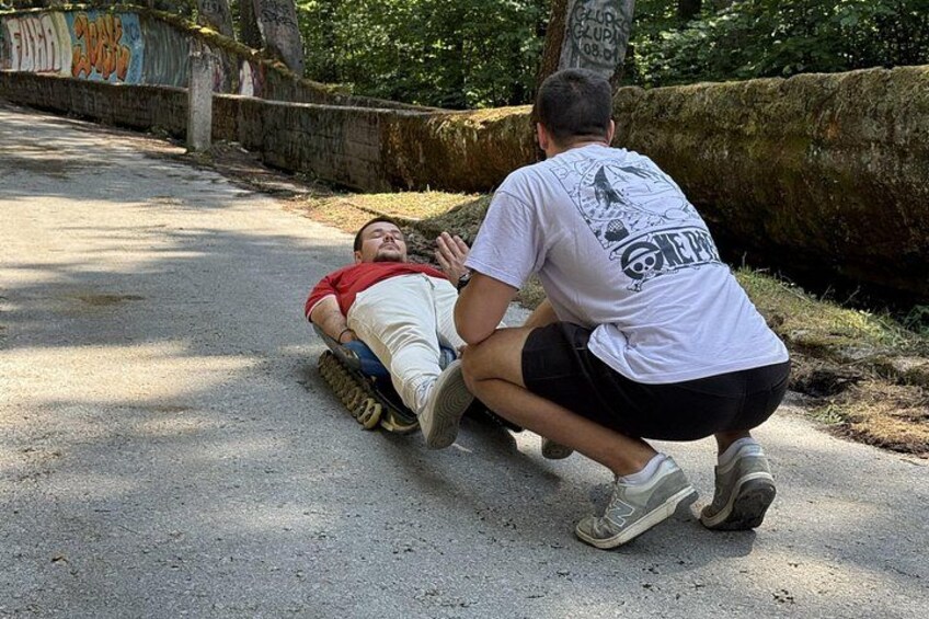 Una persona acostada en un trineo, empujada por otra, sobre un sendero pavimentado con grafitis en las paredes y una roca grande al fondo. 2
