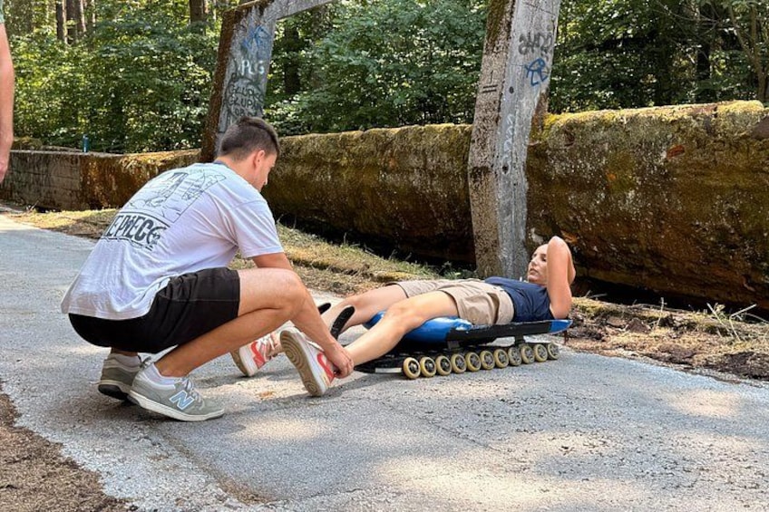 Una persona tirada en un patinete con ruedas, otra persona ayudando, y un tronco cubierto de musgo al fondo. 5
