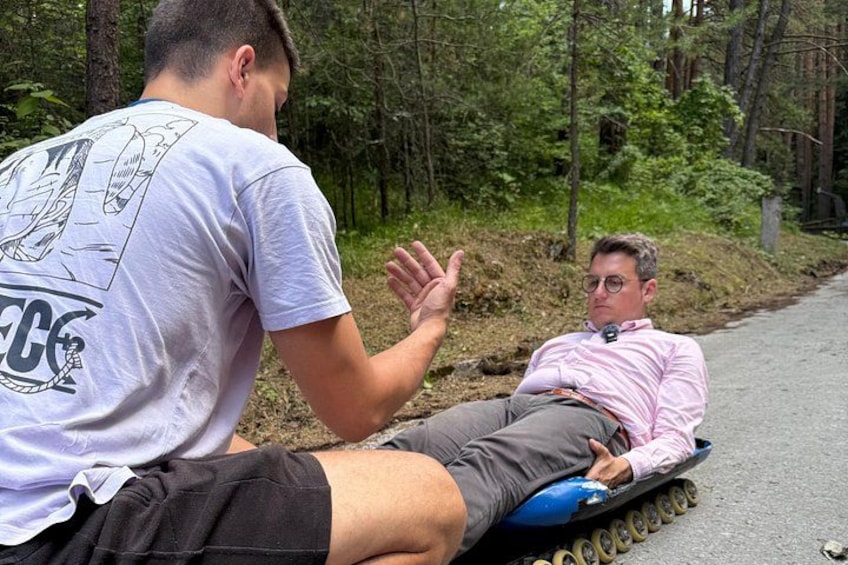 Dos personas en un trineo azul sobre un camino pavimentado, rodeado de árboles. 3