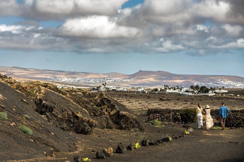 A volcanic landscape with rocky terrain, a few people walking, and a distant town under a cloudy sky. 1