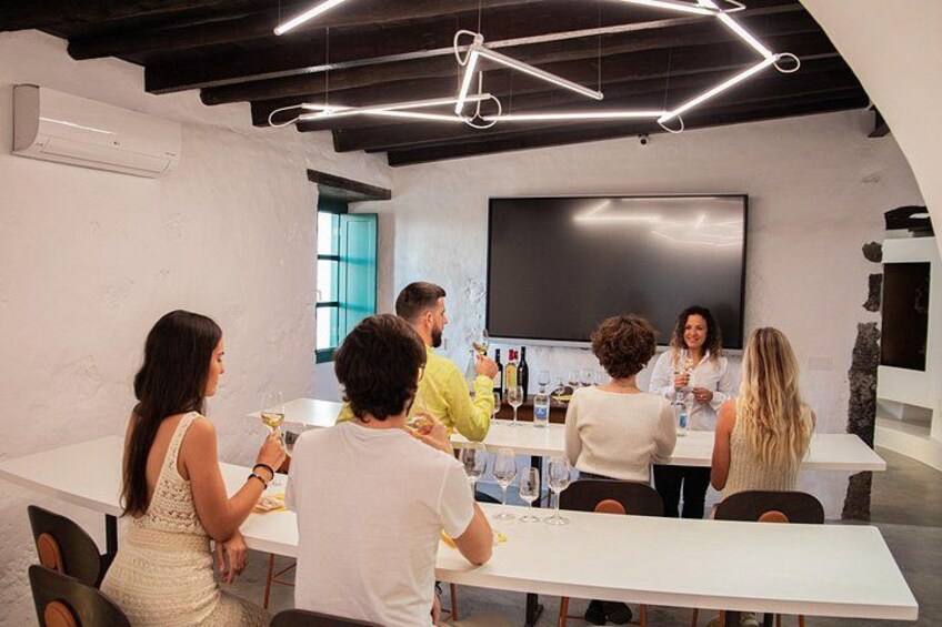 A group of people in a modern indoor setting with a long table, a flat-screen TV, and a bar area with bottles. 2