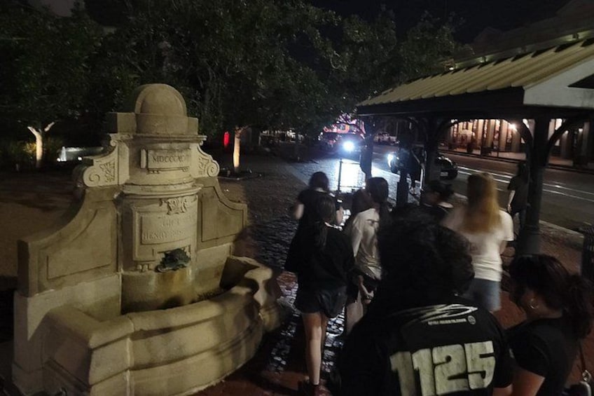 A stone fountain with inscriptions, a group of people gathered around it, and a street with vehicles in the background. 2
