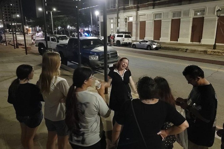 A group of people standing on a sidewalk at night, a streetlight, and a building with columns. 3