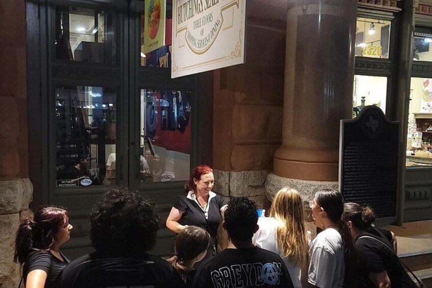 A person speaking to a crowd outside a building with large windows and a sign above. 2