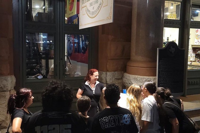 A woman speaking to a crowd outside a building with large windows and a sign above. 4