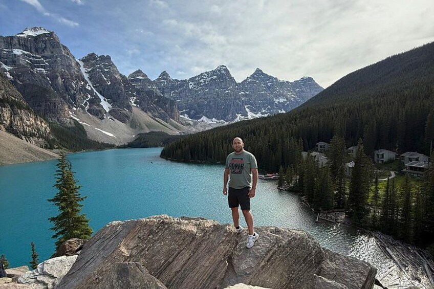 A person standing on a rocky outcrop overlooking a turquoise lake with mountains in the background. 1