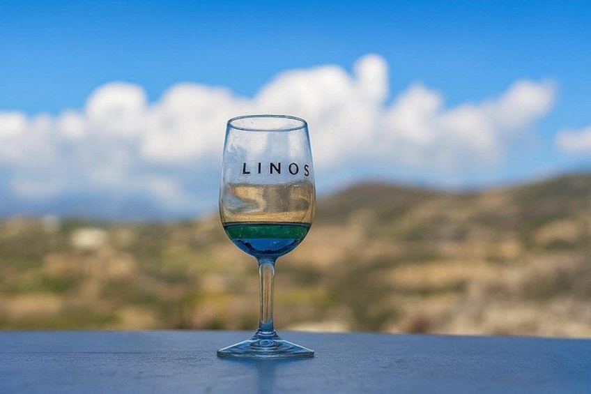 A glass of white wine with the word 'LINOS' on it, placed on a blue surface with a blurred landscape in the background. 3