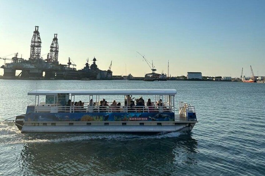 A boat with passengers, a large industrial structure in the background, and calm water. 2