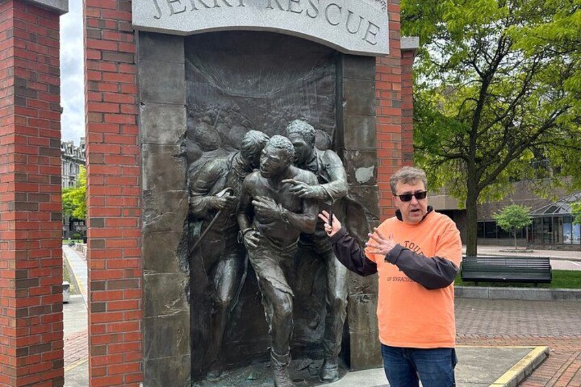 A man in an orange shirt standing in front of a sculpture titled 'JERKES RESCUE' with a relief of figures in a dramatic scene. 3