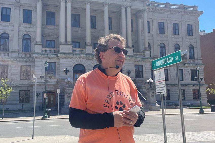A person standing in front of a building with columns, wearing an orange shirt with text, and holding a piece of paper. 2