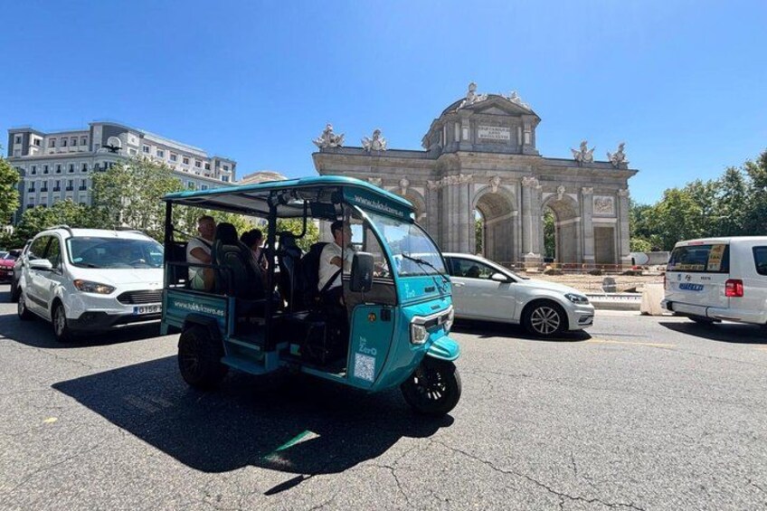 A turquoise tuk-tuk with passengers, parked in a lot with a large historical monument in the background. 3