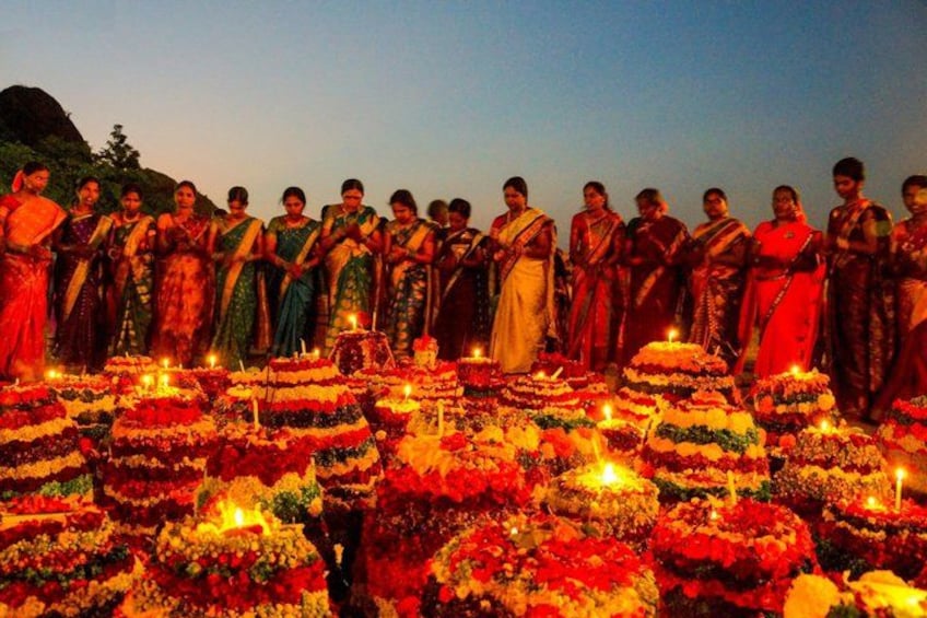 A group of women in traditional attire standing in a line, with multiple tiered floral arrangements and candles in the foreground. 2