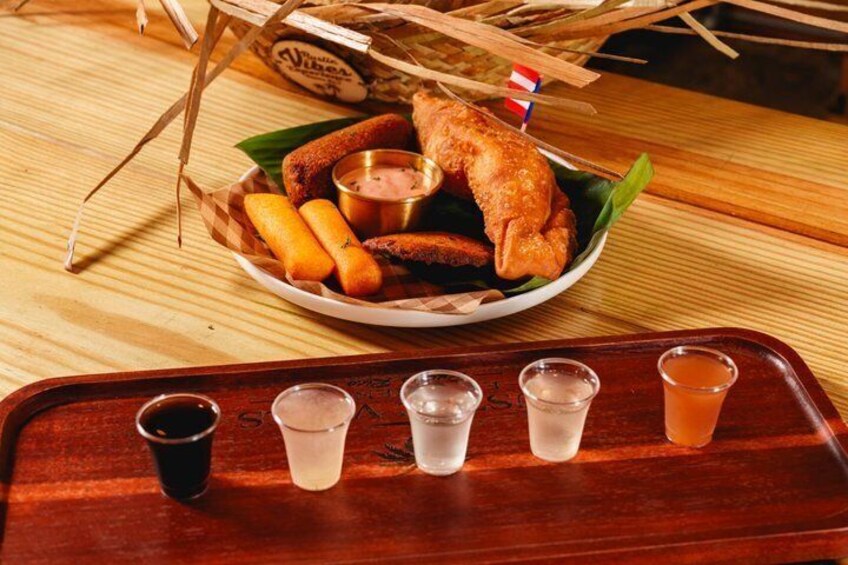 A wooden table with a tray containing a plate of fried chicken, corn dogs, and a dipping sauce, along with five shot glasses of beverages. 3