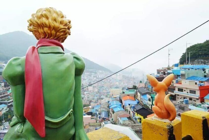 Una persona con chaqueta verde y bufanda roja observa un paisaje urbano con edificios coloridos y una montaña al fondo. 4
