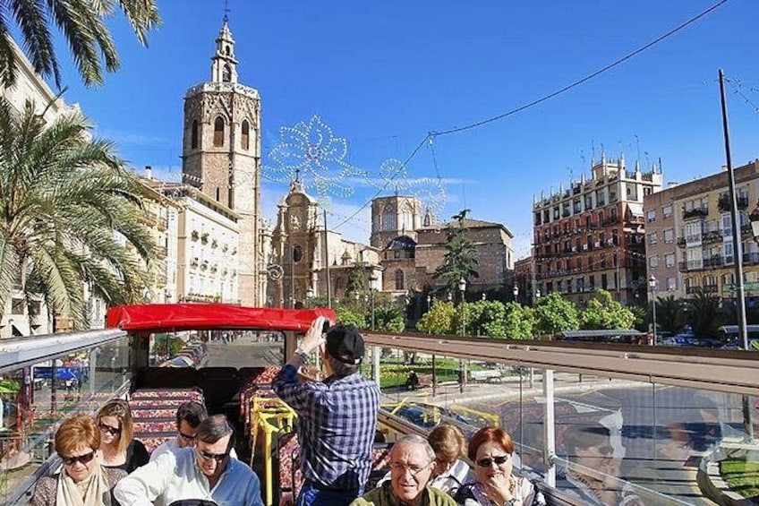 A group of people on a bus with a red roof, a historic building with a bell tower, and a Ferris wheel in the background. 5