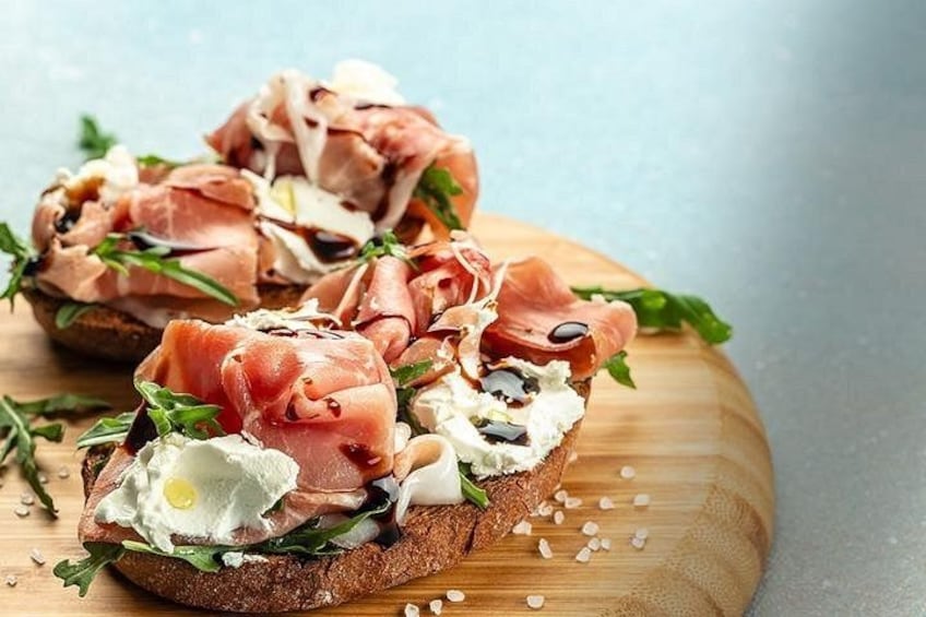 Two open-faced sandwiches with prosciutto, cheese, and greens on a wooden board. 2
