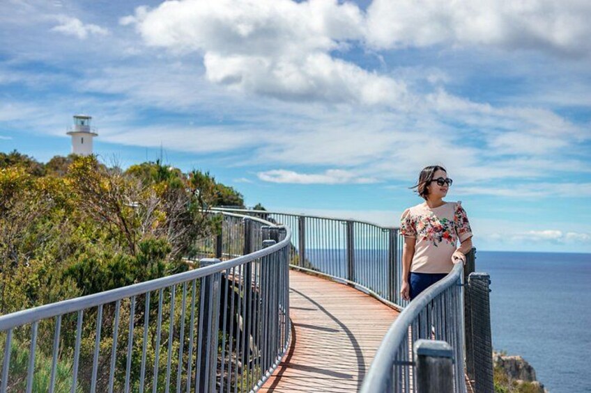 Une personne se tient sur une promenade en bois dotée de rampes métalliques, d’où elle peut admirer un paysage côtier avec un phare visible au loin. 2