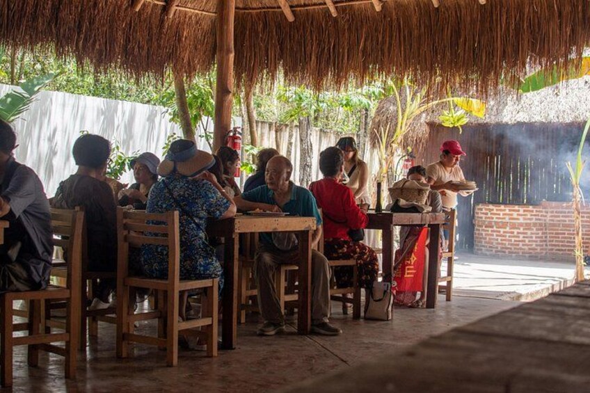 Un grupo de personas sentadas en mesas de madera bajo un techo de paja, conversando. 2