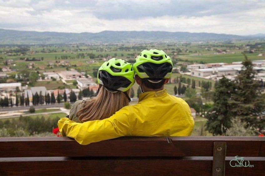 Dos personas con chalecos y cascos amarillos, sentadas en un banco con vista a un paisaje de campos y árboles. 2