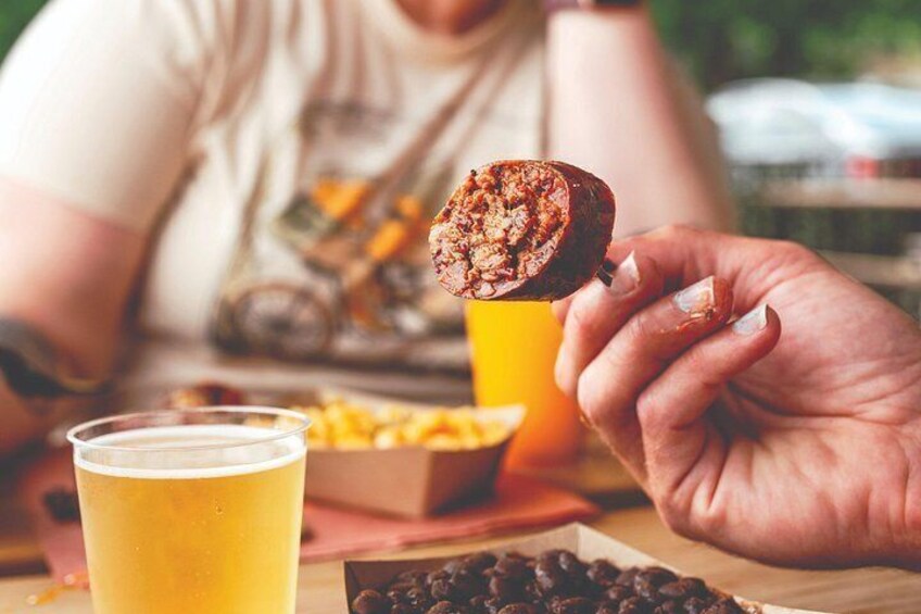 A person holding a sausage, a glass of beer, and a box of snacks at a table. 1