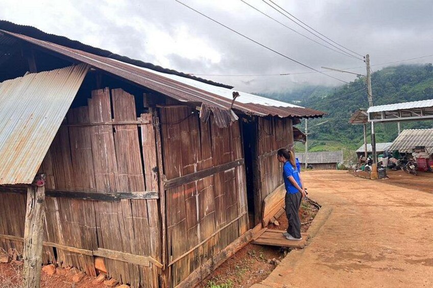 A rustic wooden structure with a corrugated metal roof, a person standing at the entrance, and a dirt road leading to a hilly landscape in the background. 2
