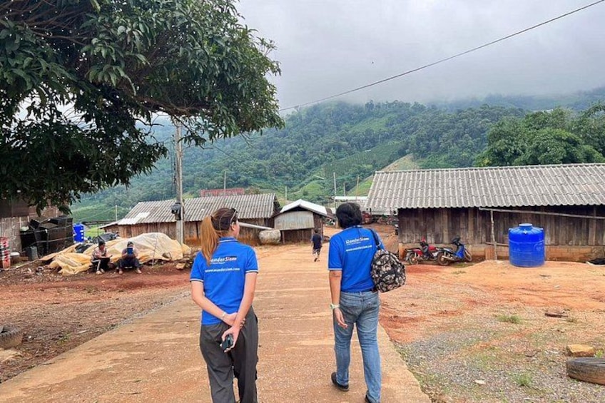 Two people in blue shirts walking on a dirt path, with buildings and a mountainous landscape in the background. 3