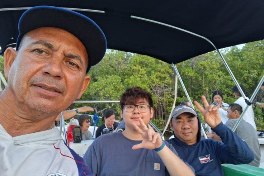 A group of people on a boat with a canopy, trees in the background, and a person making a peace sign. 2
