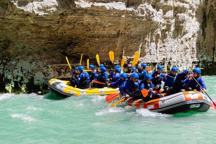 A group of people in blue helmets and orange and white rafts navigating through a river with rocky cliffs in the background. 4