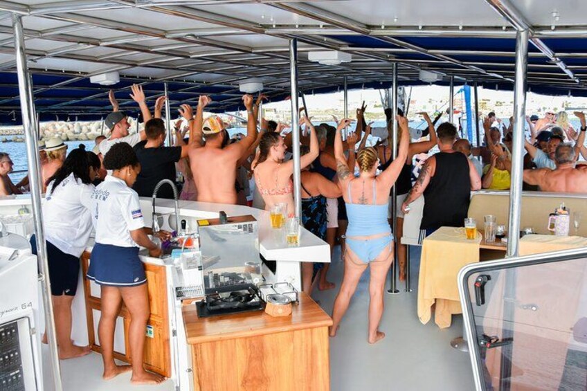 A group of people on a boat, some raising their hands, others seated at tables with drinks. 2