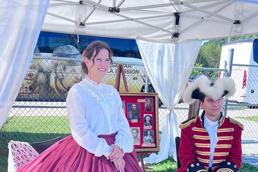 A person in a white blouse and red skirt, another in a red military uniform with a white hat, and a framed picture with photos in the background. 2