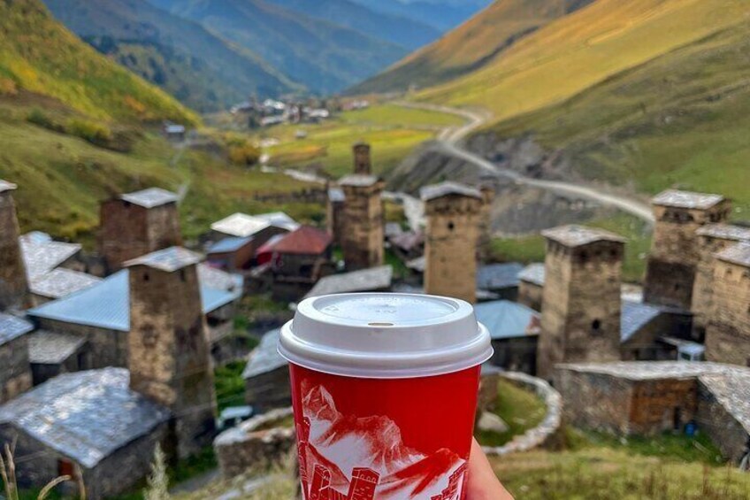 A hand holding a red coffee cup with a white design, overlooking a village with stone buildings and a winding road. 2