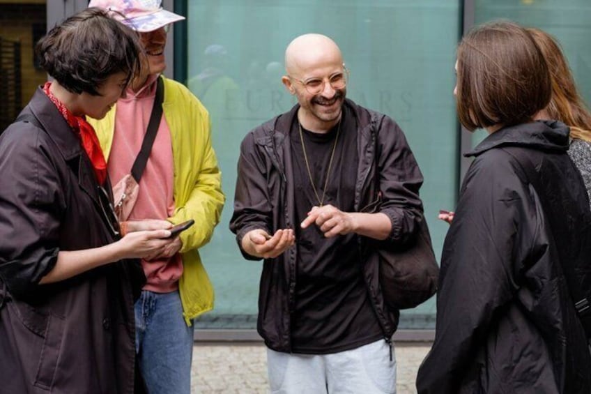 Four people standing on a sidewalk, one person holding a phone, and a building with a sign in the background. 2